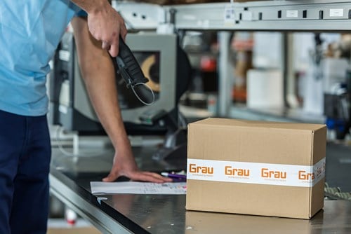 A worker scans documents at a packing station beside a sealed box labeled “Grau,”
