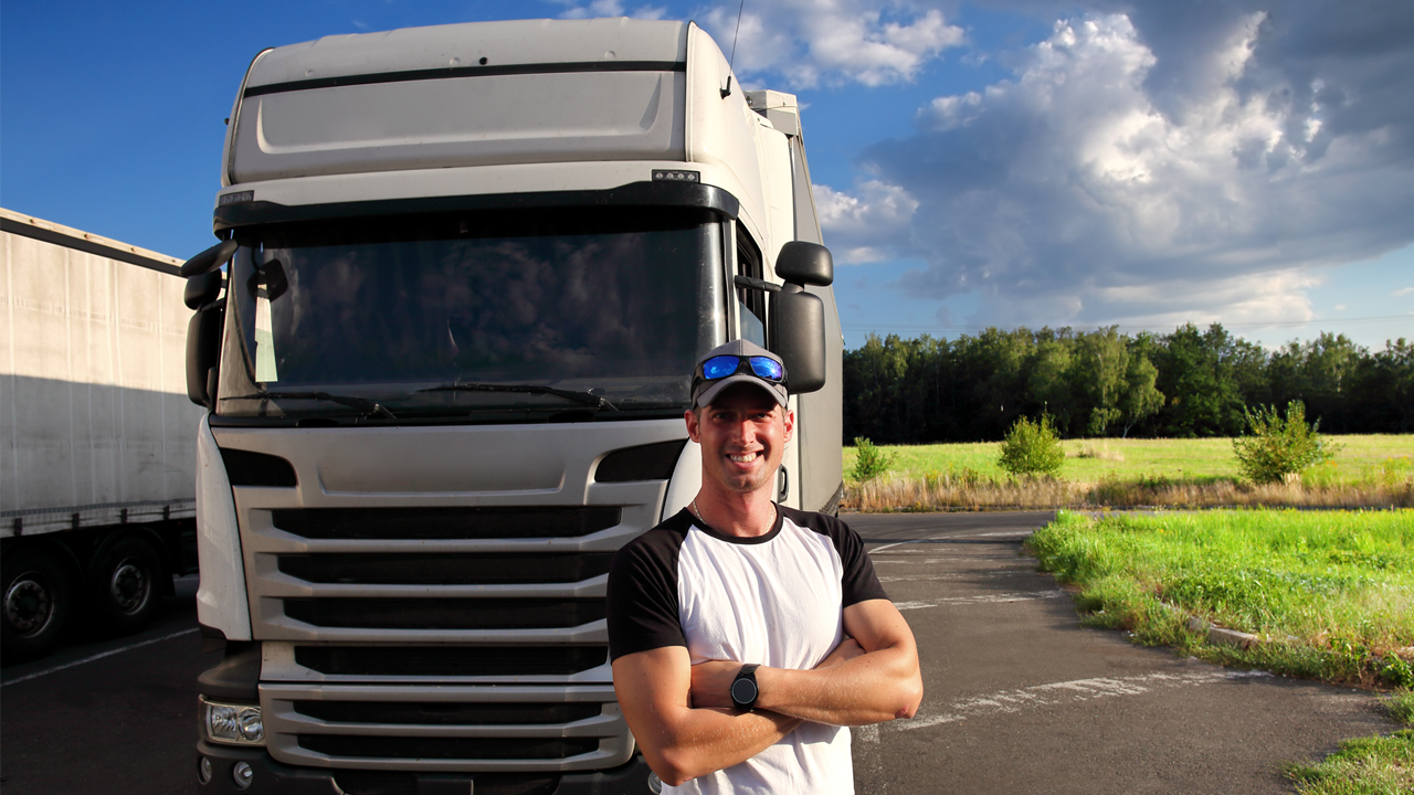 A smiling driver stands confidently with folded arms in front of a trailer