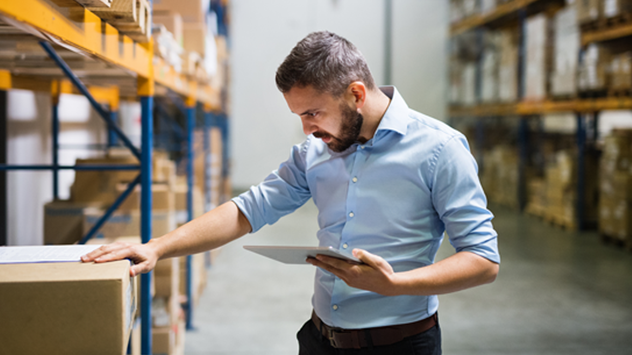 Warehouse worker inspects inventory with a tablet in hand, checking labels on stacked boxes in a storage aisle.